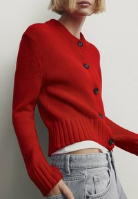 Ljus person med blont hår som bär en röd knäppt stickad kofta över en vit topp och ljusgrå jeans, stående mot en enfärgad bakgrund.