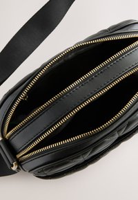Sort lædertaske med quiltet design, udstyret med flere lynlåslommer og guldfarvet hardware. Remmen er lavet af stof.