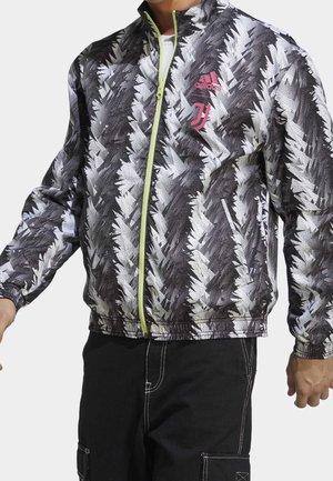 Osoba nosząca czarną, białą i szarą kurtkę Adidas w wzory z neonowo zielonym zamkiem i różowym logo Juventusu na piersi, w połączeniu z czarnymi spodniami.