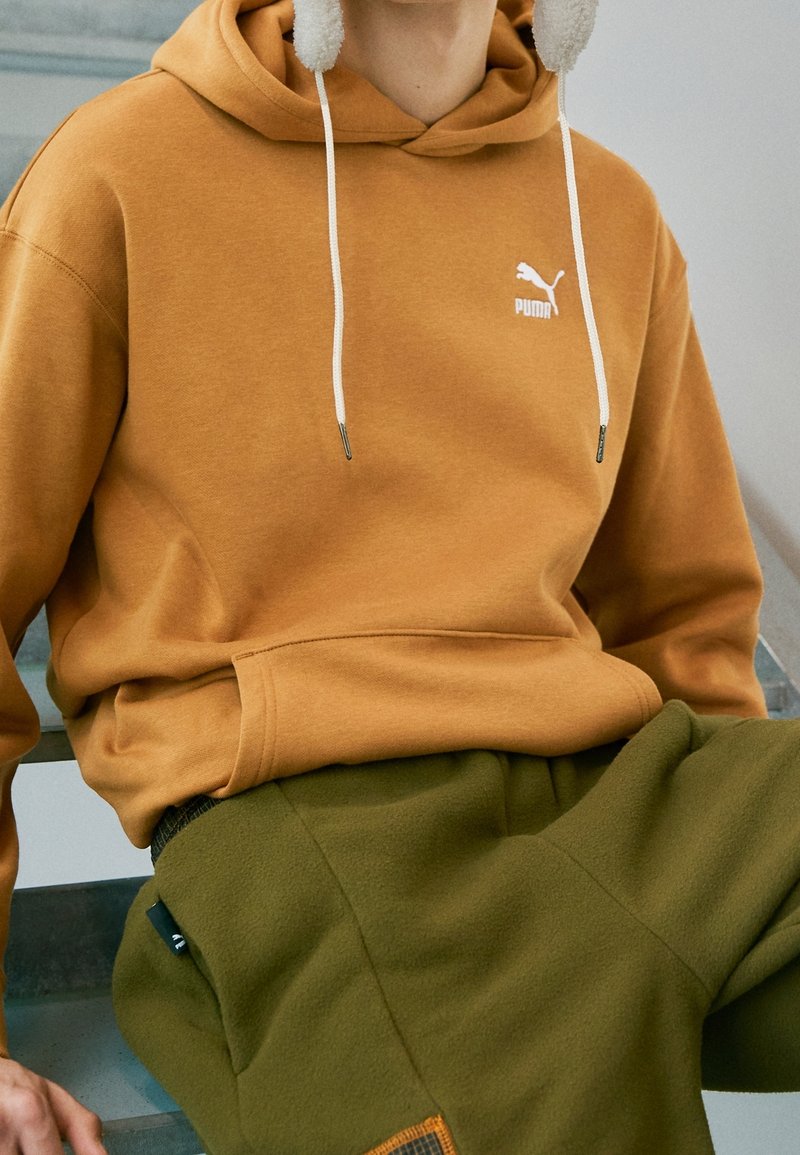 Felpa con cappuccio del marchio Puma di colore senape con lacci; indossata con pantaloni in pile verde oliva con dettagli e tasche.