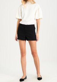 Blouse blanche à manches courtes avec des poignets bouffants associée à un short noir taille haute. Modèle portant des ballerines noires, se tenant sur un fond clair.