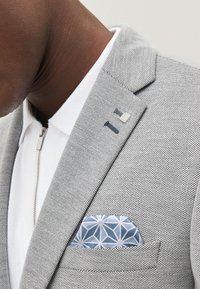 Blazer gris clair en tissu texturé, avec une chemise blanche en dessous et un mouchoir de poche bleu à motifs glissé dans la poche poitrine.