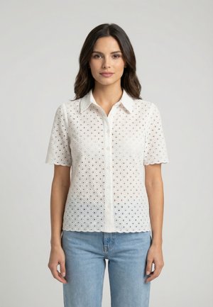 Femme aux longs cheveux foncés portant une chemise blanche à manches courtes avec œillets et un jean bleu clair, debout devant un fond neutre.
