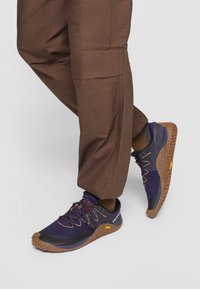 Scarpe da ginnastica viola scuro con tomaia in rete testurizzata, suole in gomma marrone e lacci arancioni a contrasto, abbinate a pantaloni cargo marroni.