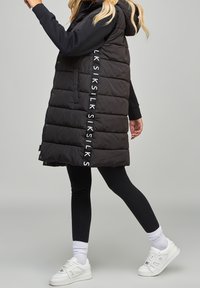 Black padded vest with a hood, featuring vertical white branding on the side. Worn over black leggings and white sneakers, with ribbed socks.