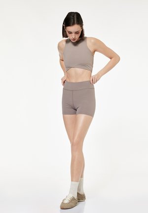 Vrouw die taupekleurige trainingsshorts met hoge taille verstelt, draagt een bijpassend mouwloos cropped topje, witte sokken en beige sneakers, staand op een witte achtergrond.