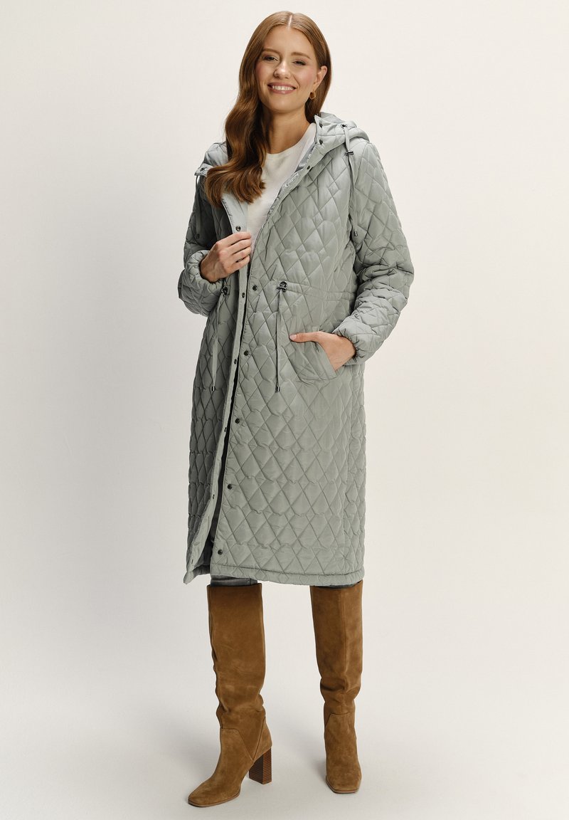 Gep quilteter langer Mantel in Hellgrau mit Kapuze, Fronttaschen und Druckknopfverschluss. Kombiniert mit hellbraunen kniehohen Stiefeln. Glatte Textur.