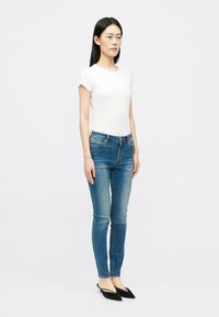 Γυναίκα που στέκεται με λευκή κοντομάνικη μπλούζα, μπλε εφαρμοστά τζιν και μαύρες γόβες slingback με μυτερές μύτες, μπροστά από μια λευκή επίπεδη επιφάνεια.