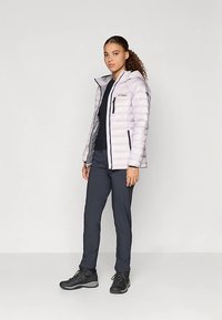 Kvinne står iført en lys lilla hettejakke, svart skjorte, mørke bukser og svarte tursko mot en lys, ensfarget bakgrunn.