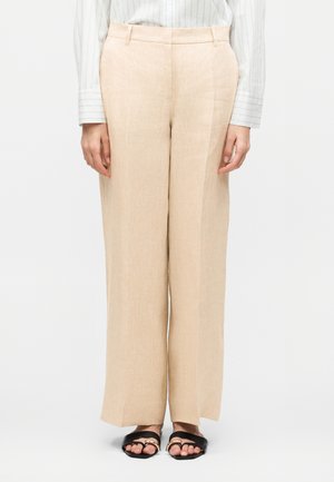 Μπεζ παντελόνια με φαρδιά μπατζάκια που φοριούνται με λευκό ριγέ πουκάμισο και μαύρα φλατ σανδάλια με χρυσές λεπτομέρειες.