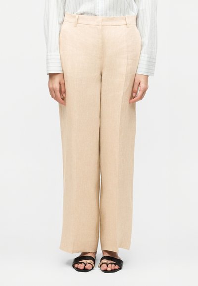 Pantalon beige à jambes larges porté avec une chemise blanche rayée et des sandales plates noires avec des accents dorés.