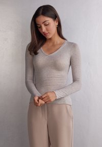Top grigio a maniche lunghe con scollatura a V, realizzato in materiale morbido, con un design aderente e una texture liscia, abbinato a pantaloni beige.