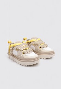 Zapatillas beige y blancas con suelas gruesas y cordones contrastantes en amarillo y beige, situadas una al lado de la otra sobre un fondo blanco.