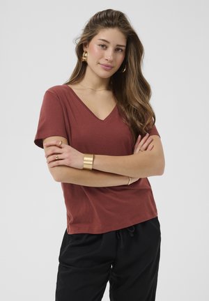 Jeune femme aux longs cheveux bruns portant un shirt à col en V couleur rouille, un pantalon noir et des bijoux dorés, debout les bras croisés.