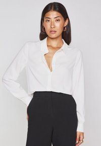 Blusa bianca a maniche lunghe con colletto a punta e chiusura con bottoni, abbinata a pantaloni neri a vita alta. Tessuto morbido, taglio classico.