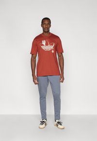 Κόκκινο βαμβακερό T-shirt με λευκό γραφικό σχέδιο και κείμενο, συνδυασμένο με γκρι φόρμα με πλάγιες ρίγες και μπεζ αθλητικά παπούτσια.