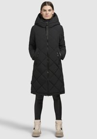Černý prošívaný dlouhý kabát s kapucí, zapínáním na zip a bočními kapsami. Hladká tkanina s geometrickým diamantovým vzorem. V kombinaci s černými legínami a světlými botami.