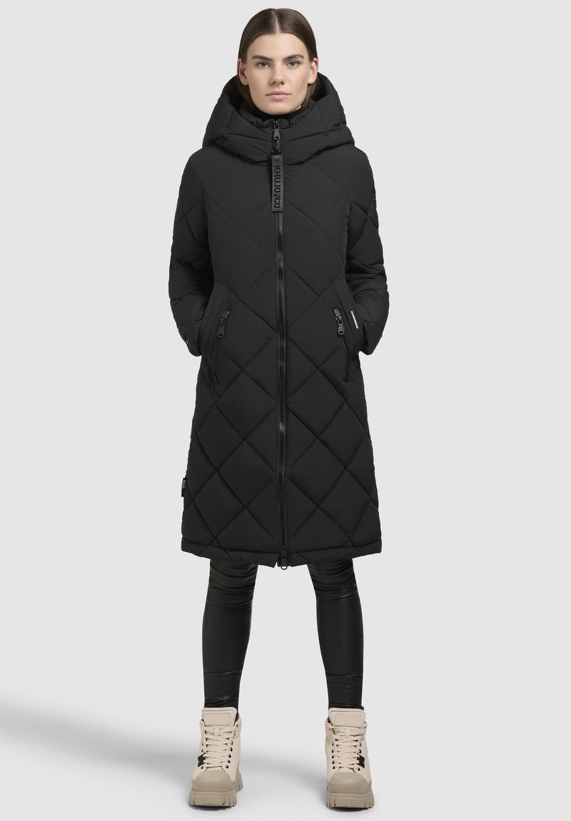 Černý prošívaný dlouhý kabát s kapucí, zapínáním na zip a bočními kapsami. Hladká tkanina s geometrickým diamantovým vzorem. V kombinaci s černými legínami a světlými botami.