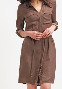 Hnědé šaty s krátkým rukávem, dvěma náprsními kapsami, knoflíkovým zapínáním vpředu a stahovacími šňůrkami v pase. Hladký materiál s mírným leskem, po kolena.