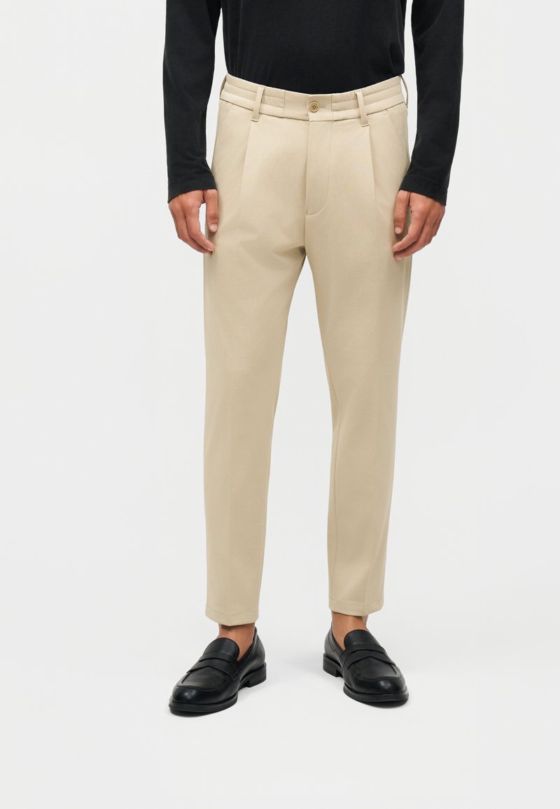 Mann trägt beige, maßgeschneiderte Hose mit Gürtelschlaufen, Falten, schwarzen Slippern und einem langärmligen schwarzen Hemd, steht vor weißem Hintergrund.