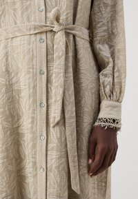 Beige textured dress with a leaf pattern, tie waist, and button front. Features long sleeves with fringed cuffs and smooth fabric.