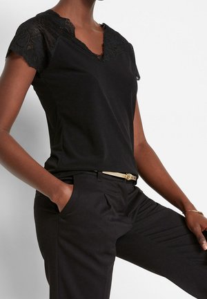 Femme portant un haut noir en dentelle à manches courtes avec une ceinture fine en or, un pantalon noir, une main dans la poche, debout devant un fond blanc.