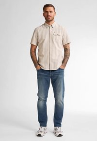 Kurzärmeliges, hellbeiges gestreiftes Hemd mit Knopfleiste und Taschen-Detail, kombiniert mit blauen Jeans und weißen Turnschuhen.