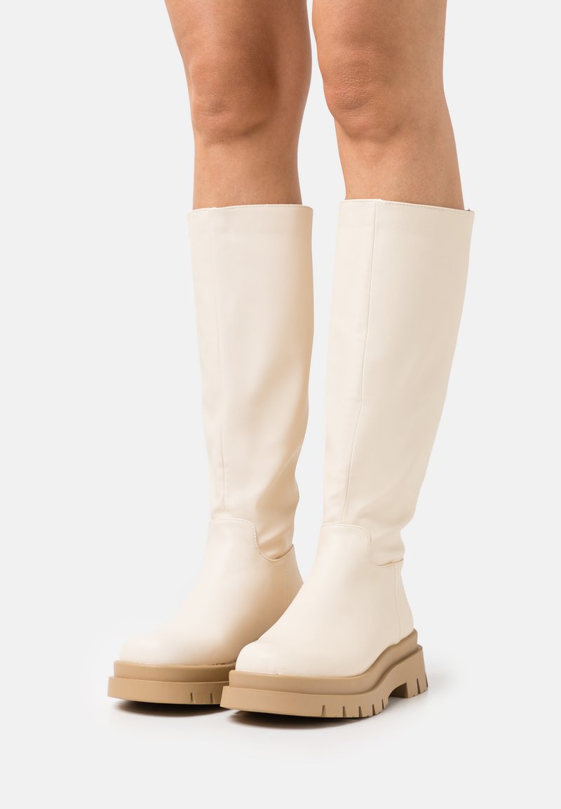 Botas brancas até ao joelho, feitas de um material liso, com uma sola bege chunky e biqueira arredondada. Apresentam um design simples e aerodinâmico.