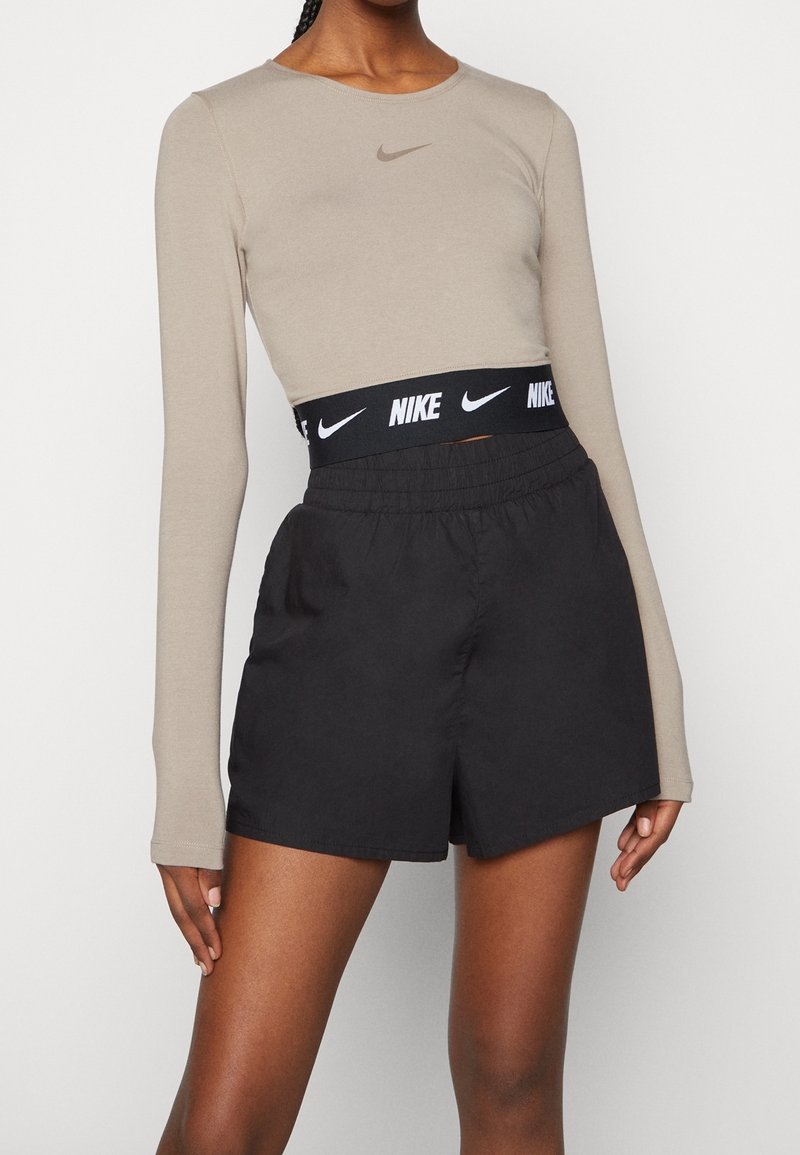 Maglietta a maniche lunghe beige con logo Nike, abbinata a pantaloni corti neri con una cintura spessa, anch'essi marchiati Nike in bianco.