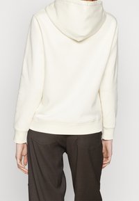 Sudadera con capucha de color crema con capucha, puños y dobladillo acanalados, vista desde atrás. Combinada con pantalones marrón oscuro. Textura suave.