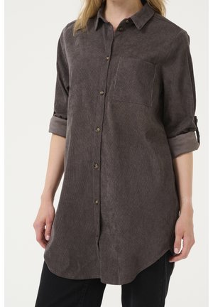 Camisa de pana de manga larga en color marrón oscuro con cierre de botones, bolsillo en el pecho y puños enrollados. Textura suave y dobladillo curvado.