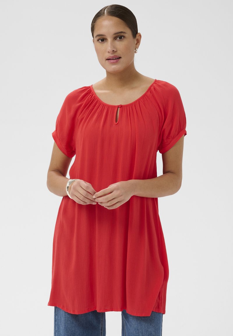 Vrouw met bruin haar in een lage knot, gekleed in een losse rode korte mouw tuniek over een blauwe spijkerbroek, staand tegen een effen lichte achtergrond.