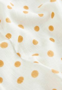 Tissu blanc avec un tissage texturé et des pois orange espacés uniformément, présenté avec des plis doux et de légères ombres.