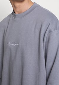 Sweat-shirt gris en tissu doux, avec un col ras du cou côtelé et un discret détail brodé sur la poitrine.