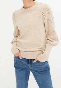 Beige stickad tröja med mönstrade puffärmar i kombination med blå denimjeans, modellens händer vilande i jeanfickorna.