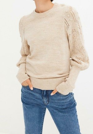 Beige gebreide trui met patroon mouwen en een pof, gecombineerd met blauwe jeans, met de handen van het model in de broekzakken.