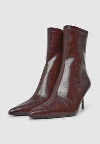 Stivali alla caviglia con punta affusolata in un lucido marrone scuro, con tacchi alti e sottili, realizzati in pelle dalla superficie liscia su uno sfondo semplice.