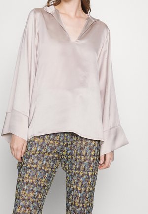 Femme portant un chemisier ample en satin rose clair avec de larges poignets et un pantalon multicolore à motifs sur fond uni.