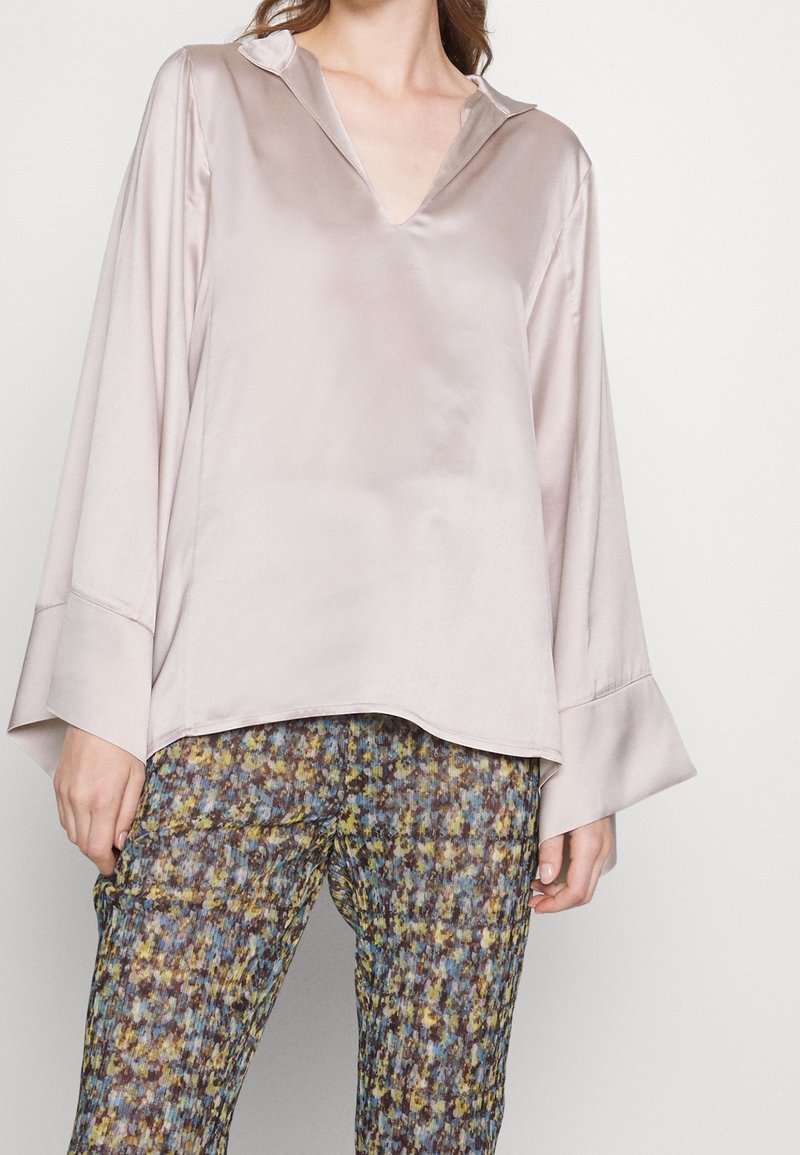 Femme portant un chemisier ample en satin rose clair avec de larges poignets et un pantalon multicolore à motifs sur fond uni.