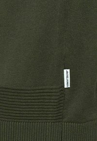 Pull vert foncé en tricot avec une texture côtelée en bas et une petite étiquette blanche "JACK & JONES" sur le côté.