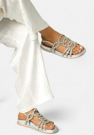 Pieds portant des sandales argentées avec des brides tressées scintillantes, associées à un pantalon blanc texturé et ample sur un fond uni.