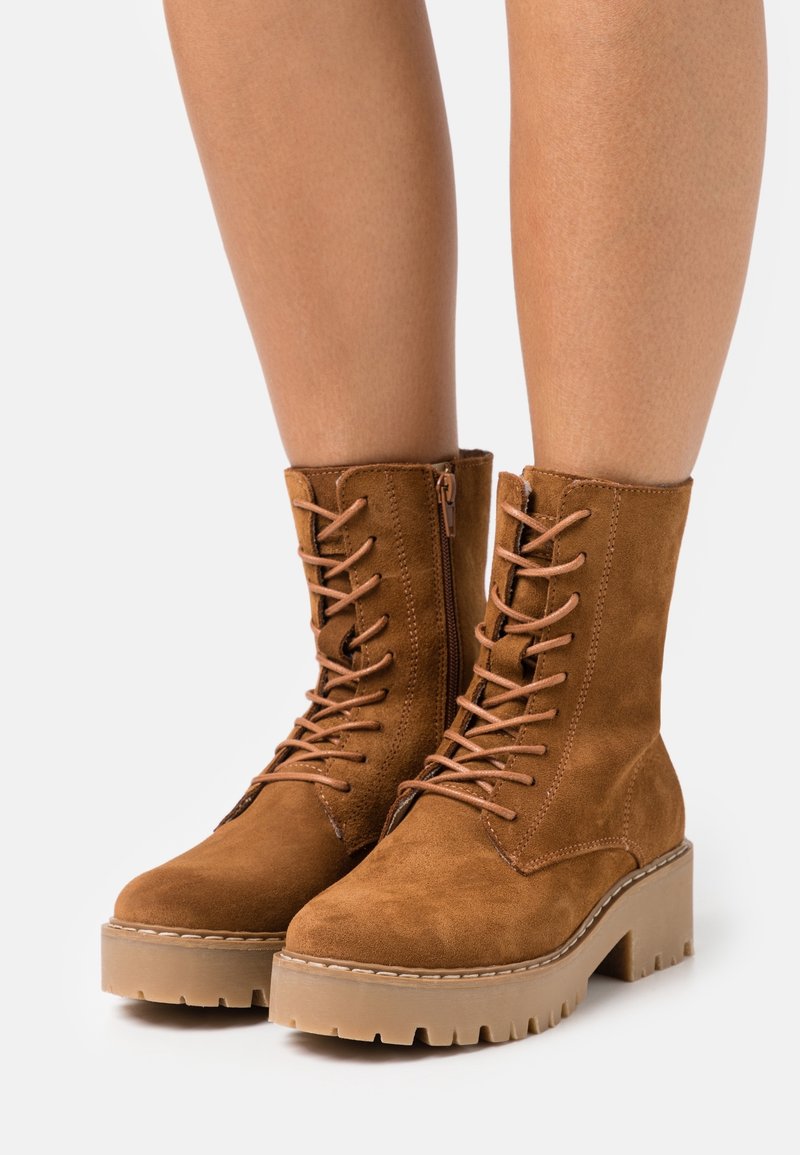 Bottes montantes en daim marron à lacets avec semelles en caoutchouc épaisses portées sur des jambes nues sur un fond blanc.