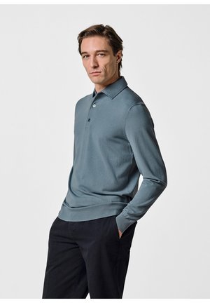 Uomo con polo a maniche lunghe blu e pantaloni neri, in piedi con le mani in tasca contro uno sfondo bianco uniforme.