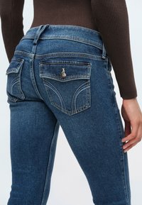 Detailný záber na osobu nosiacu modré džínsy z denimu s gombíkmi na zadných vreckách a hnedý rebrovaný dlhý rukáv, ruka spočíva pri stehne.