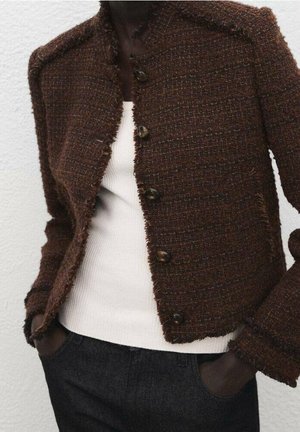 Personne portant une veste marron texturée boutonnée sur un pull blanc côtelé, les mains dans les poches de pantalons foncés.