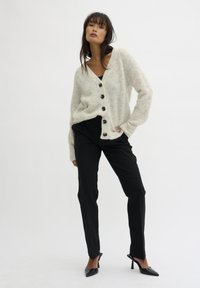 Svetlo šedý, voľný cardigan s čiernymi gombíkmi, nosený cez čierny top a čierne nohavice. Tkanina vyzerá jemne a textúrne. Vysoké podpätky dopĺňajú outfit.