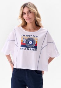 T-shirt blanc court avec un col rond, des manches courtes, des coutures bleues et un imprimé graphique représentant un disque vinyle et du texte.