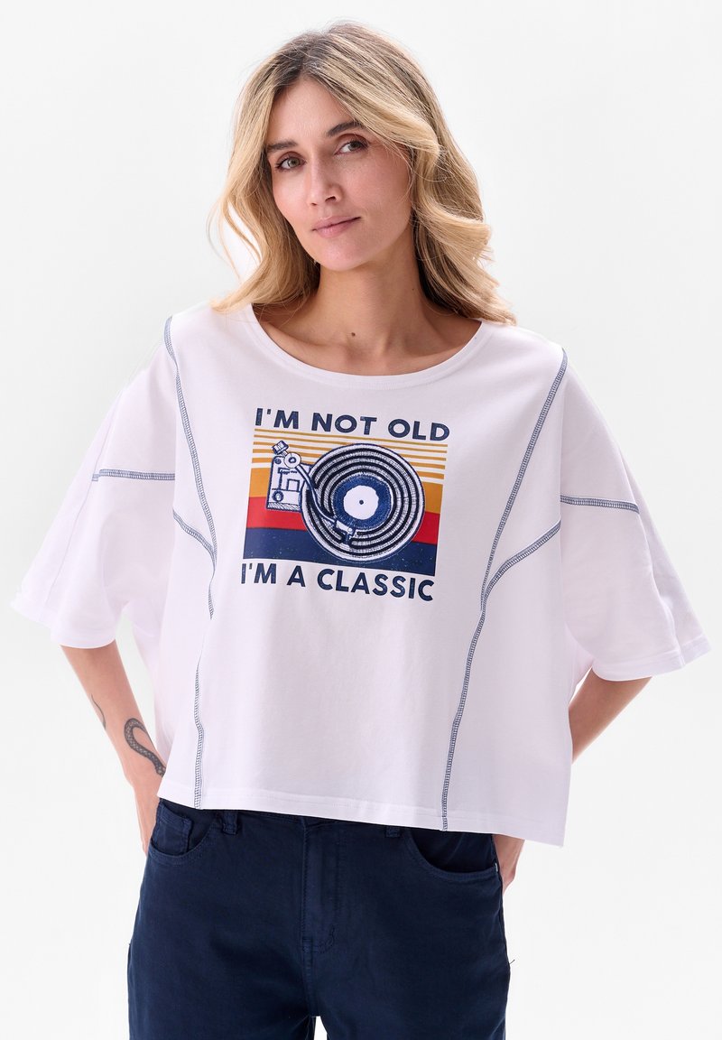 T-shirt blanc court avec un col rond, des manches courtes, des coutures bleues et un imprimé graphique représentant un disque vinyle et du texte.