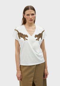 T-shirt bianco in cotone con scollo a V e maniche corte. Presenta motivi leopardati metallici in oro e nero sulle spalle. Texture liscia.