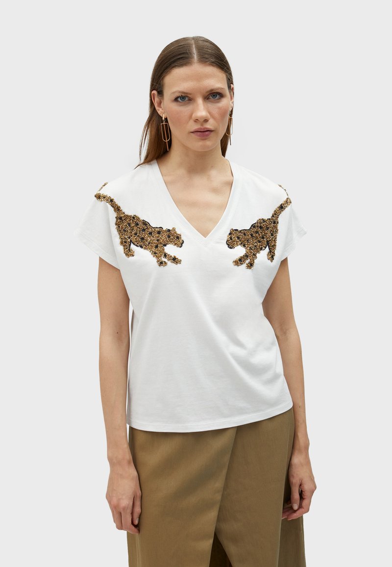 T-shirt bianco in cotone con scollo a V e maniche corte. Presenta motivi leopardati metallici in oro e nero sulle spalle. Texture liscia.
